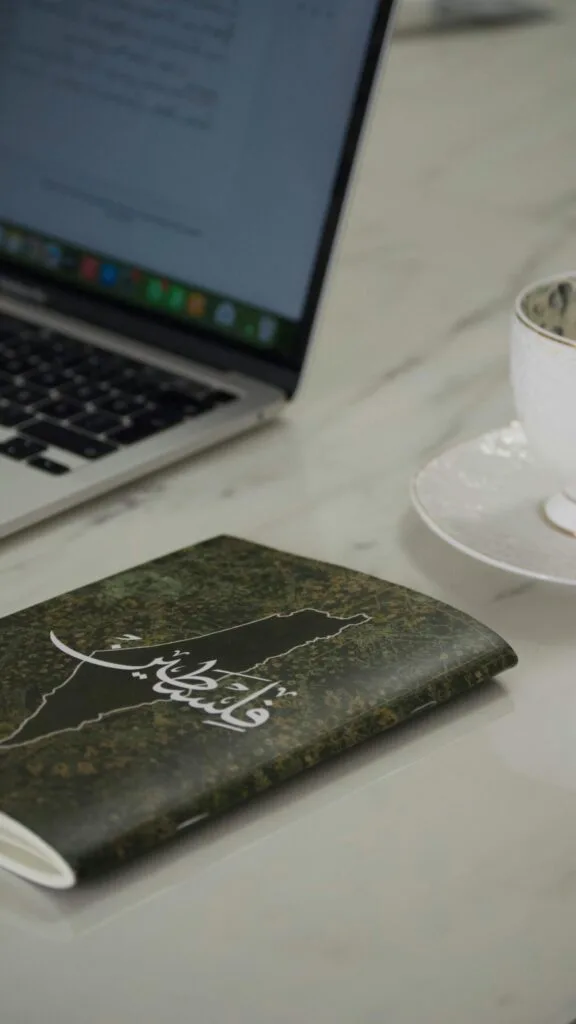 Carnet à couverture verte avec une inscription en arabe posé sur une table près d’un ordinateur portable et d’une tasse blanche.