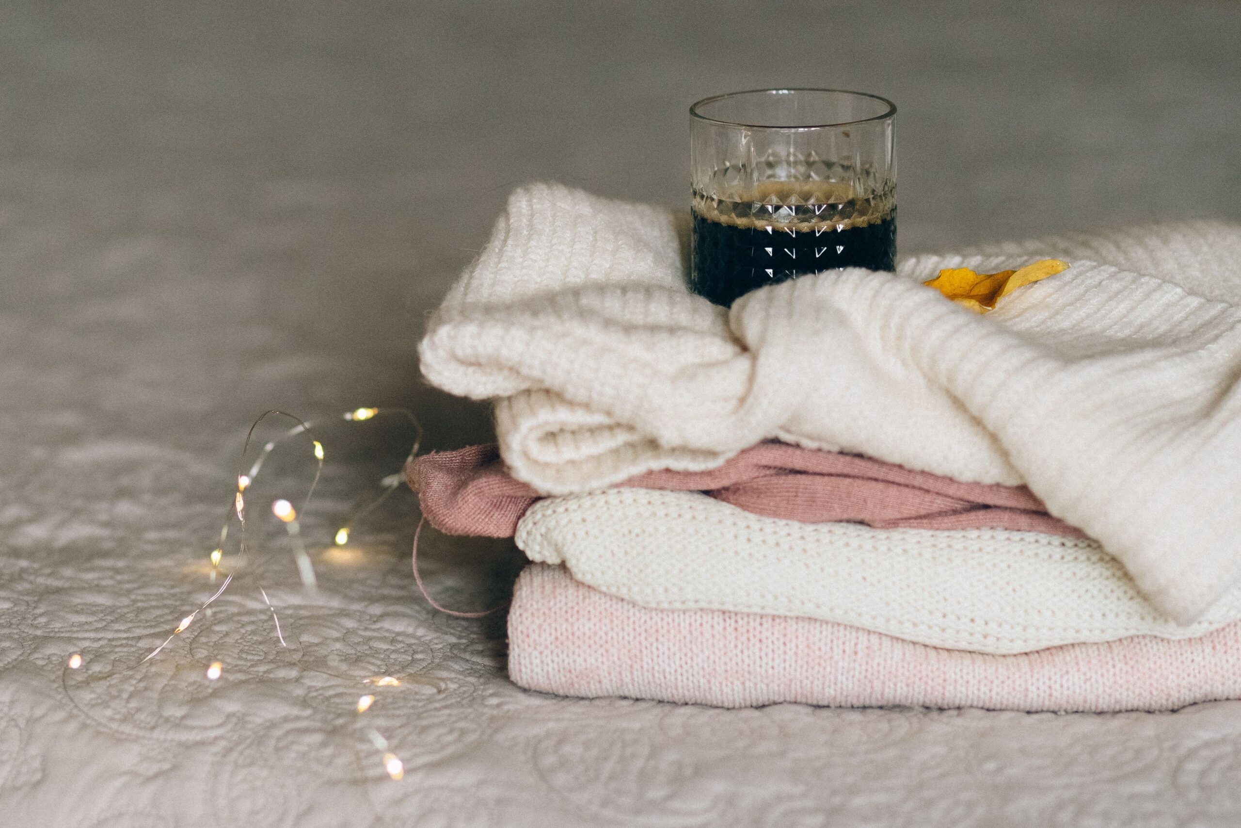 Pile de pulls en tricot doux empilés avec une boisson chaude et des lumières cosy évoquant le confort hivernal