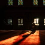 Silhouette d'une personne en prière dans une mosquée, éclairée par la lumière naturelle traversant les vitraux colorés