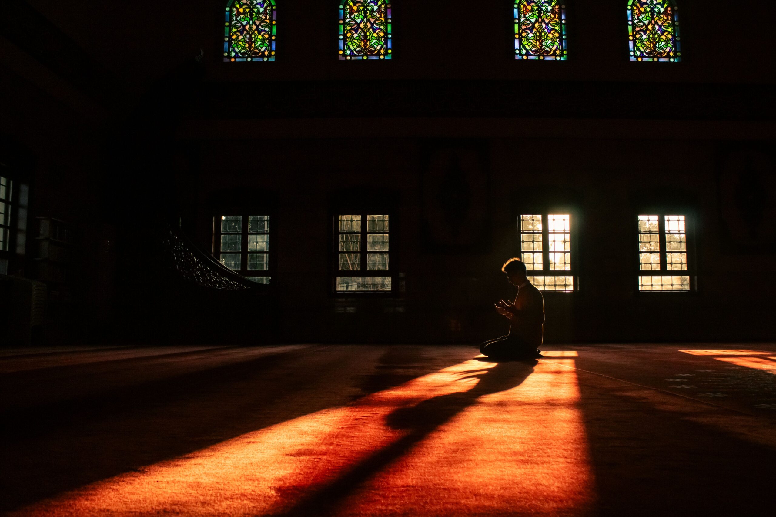 Silhouette d'une personne en prière dans une mosquée, éclairée par la lumière naturelle traversant les vitraux colorés